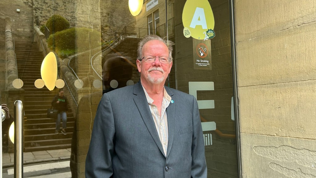 Howard Priestley smiles down the camera as he stands outside of the building where Phoenix now broadcast from. An E for 'E Mill' can be seen behind him. 